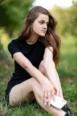 Parkta ya da bahçede yeşil çimlerde oturan ve elinde telefon tutan genç ve güzel bir kız. dikey resim