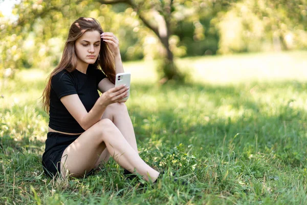 a girl from a TV station sits in a green park in the shade on a sunny day. the girl looks at the phone