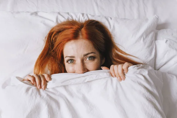 Coy young woman hiding under bed cover — Stockfoto