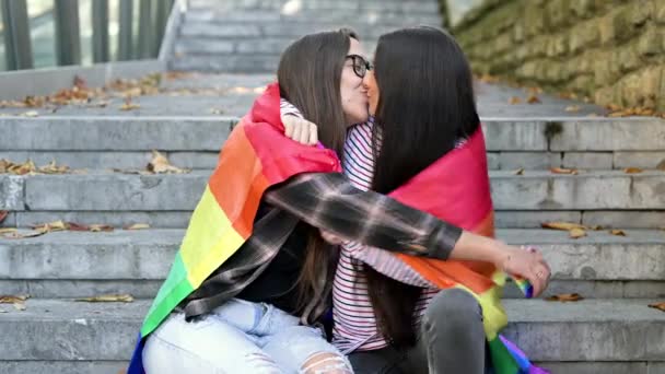 Pareja lesbiana sentada en escalones con bandera de arco iris, abrazándose y besándose en el paisaje urbano. — Vídeos de Stock