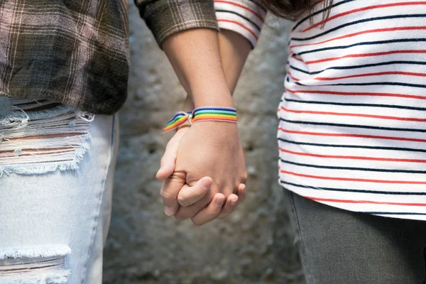 Close up de feliz casal lésbico de mãos dadas usando pulseira bandeira arco-íris, conceito de amor LGBT. — Fotografia de Stock