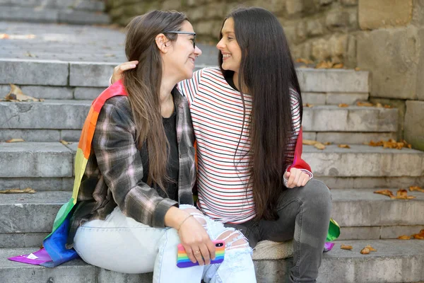 Embarazada pareja lésbica, sentado en escaleras abrazando con la bandera del arco iris en el paisaje urbano. —  Fotos de Stock