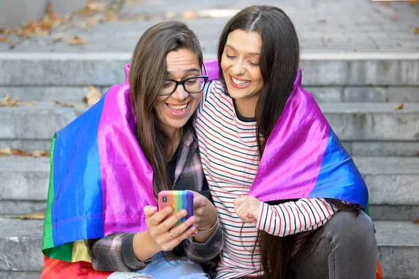 Hermosa pareja lesbiana con bandera de arco iris usando un teléfono móvil en la calle. —  Fotos de Stock