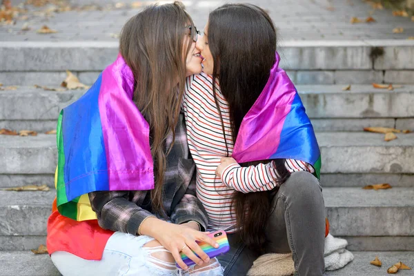 Pareja lesbiana sentada en escalones con bandera de arco iris, abrazándose y besándose en el paisaje urbano. —  Fotos de Stock