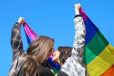 Mavi gökyüzünün altında LGBT bayrağı taşıyan sevecen lezbiyen çift.