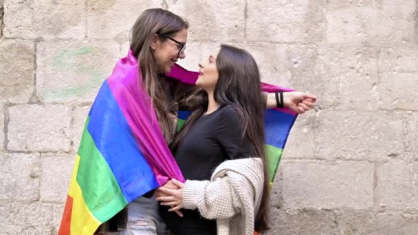 Portret ciężarnej młodej lesbijki przytulającej się podczas trzymania tęczowej flagi przed kamienną ścianą na zewnątrz — Wideo stockowe