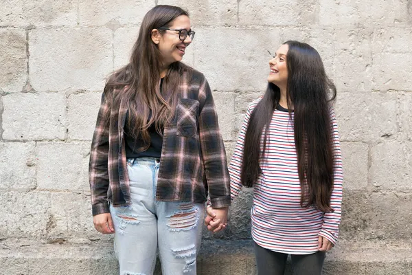 İki genç lezbiyen birbirlerini tutuyorlar ve taş bir duvarın yanında duruyorlar, gülümsüyorlar, mutlular.. — Stok fotoğraf