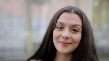 Güneşli bir günde dışarıda poz veren güzel genç kadın portresi, arka planda bulanık şehir manzarası, kameraya gülümsüyor..