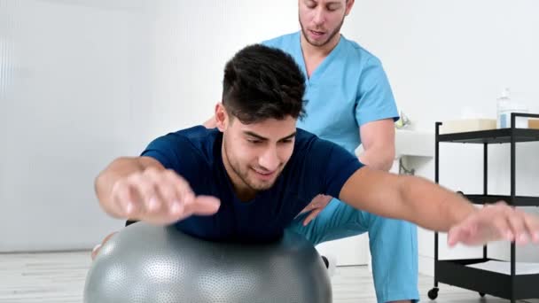 Fisioterapeuta ayuda apuesto joven paciente para hacer ejercicios de pilates con pelota. — Vídeos de Stock