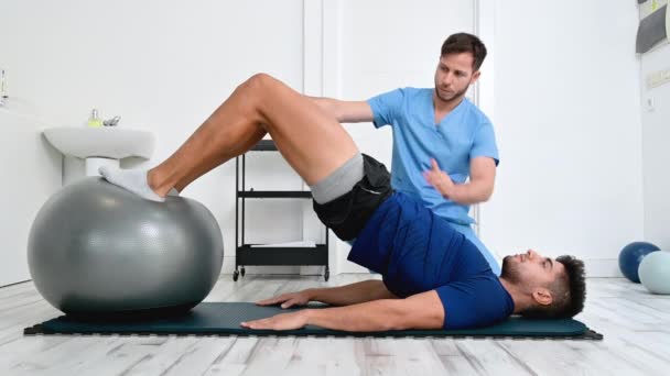 Fizioterapeuta segít jóképű fiatal beteg csinál pilates gyakorlatok labda. — Stock videók