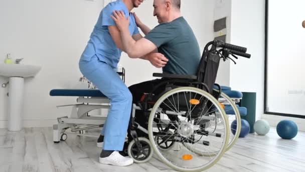 Fisioterapeuta masculino que ayuda a un paciente con una enfermedad neurológica a levantarse de una silla de ruedas en el hospital de rehabilitación. — Vídeos de Stock