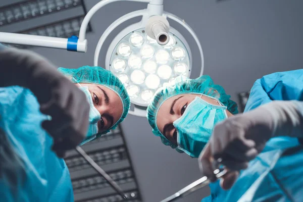 수술실에서 밝은 빛 아래서 있는 동안 전문 도구를 사용하는 의료 복을 입은 여성 외과 의사들 아래에서 — 스톡 사진