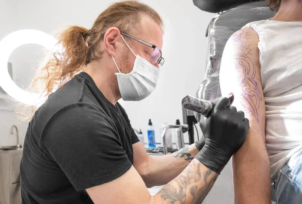 Tatuaje con su máquina tatuándose en el brazo con el diseño dibujado y usando máscara protectora contra covid 19, concepto de arte y diseño durante la nueva normalidad. — Foto de Stock