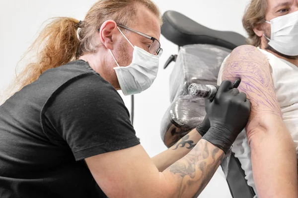 Tatuaje con su máquina tatuándose en el brazo con el diseño dibujado y usando máscara protectora contra covid 19, concepto de arte y diseño durante la nueva normalidad. — Foto de Stock