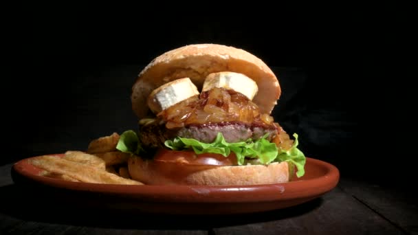 Wysokiej jakości hamburger wołowy z kozim serem, karmelizowana cebula w rustykalnej bułce z frytkami na boku. Pyszne Burger serwowane na rustykalnym talerzu na drewnianym stole — Wideo stockowe