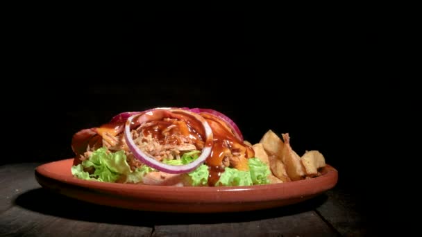 Dekat dengan seorang koki yang tidak dikenal sedang menyelesaikan burger babi yang lezat, disajikan dengan kentang goreng di atas piring berbahan kayu — Stok Video