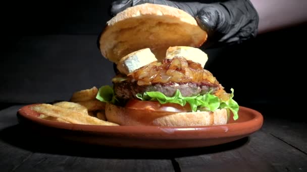 Primo piano di uno chef irriconoscibile che rifinisce un hamburger di qualità Premium con formaggio di capra e cipolle caramellate, servito con patatine fritte su un piatto rustico sul tavolo di legno — Video Stock