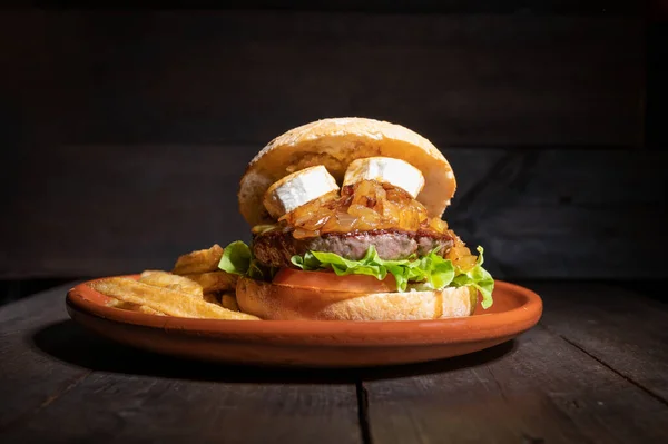 Premium quality Beef burger with goat cheese, caramelized onions in a rustic bun with french fries on the side. Yummy Burger served on a rustic plate on wooden table