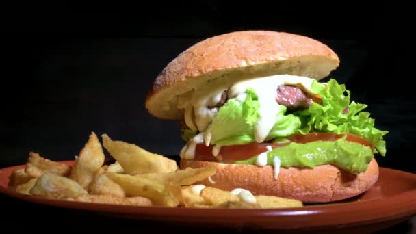 Finom ínyenc marhahús burger krémes mártással, salátával, paradicsommal és guacamole-lal, sült krumplival tálalva egy rusztikus tányéron egy fa asztalon. Lövöldözés. Kiváló minőségű 4k felvételek — Stock videók