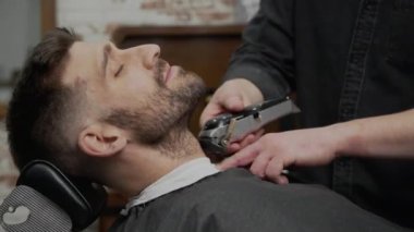 Modern berber dükkanında sakal tımar ederken genç hippi beyaz adam. Erkek saç stili. Yakışıklı adam elektrikli budayıcı ile yeni saç stili alıyor.. 