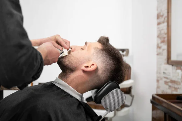 Молодой белый хипстер во время ухода за бородой в современной парикмахерской. Мужская прическа. Красивый мужчина получает новую прическу с электрическим триммером. — стоковое фото