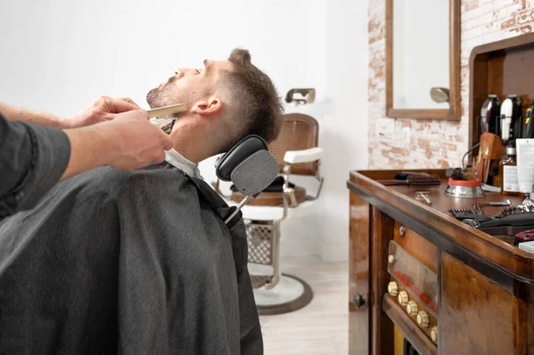 Jonge hippe blanke man tijdens baardverzorging in een moderne kapperszaak. Mens haar styling. Knappe man krijgt nieuw kapsel met elektrische trimmer. — Stockfoto