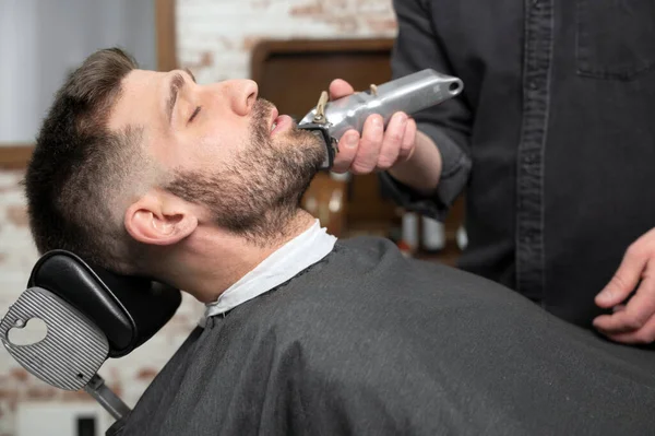 在现代理发店修整胡子时，年轻的嬉皮士白人男子。男人的发型风格。一个英俊的男人用电动理发机改头换面. — 图库照片