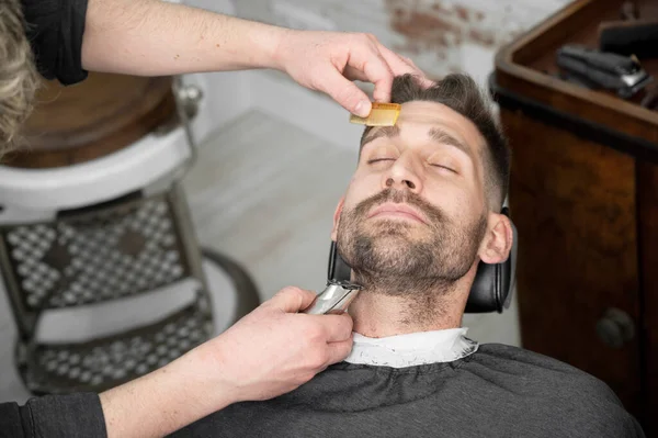 Jonge hippe blanke man tijdens baardverzorging in een moderne kapperszaak. Mens haar styling. Knappe man krijgt nieuw kapsel met elektrische trimmer. — Stockfoto