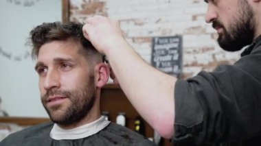 Erkek berber, berber dükkanında kırpıcıyla erkek müşterinin saçını kesiyor. Saç şekillendirme süreci. 
