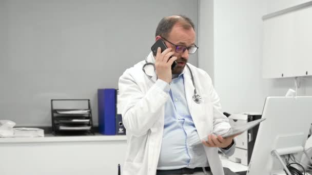 Male physician with stethoscope sitting at desk, talking on mobile phone and looking at digital tablet, discussing a patient diagnostic. — Vídeo de stock
