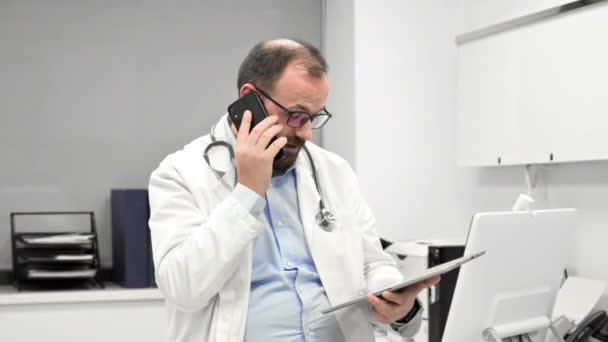 Male physician with stethoscope sitting at desk, talking on mobile phone and looking at digital tablet, discussing a patient diagnostic. — Vídeo de stock