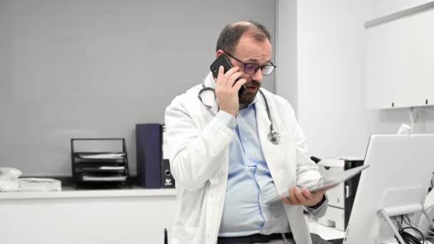 Mužský lékař se stetoskopem sedí za stolem, mluví na mobilním telefonu a dívá se na digitální tablet, diskutuje o diagnostice pacienta. — Stock video