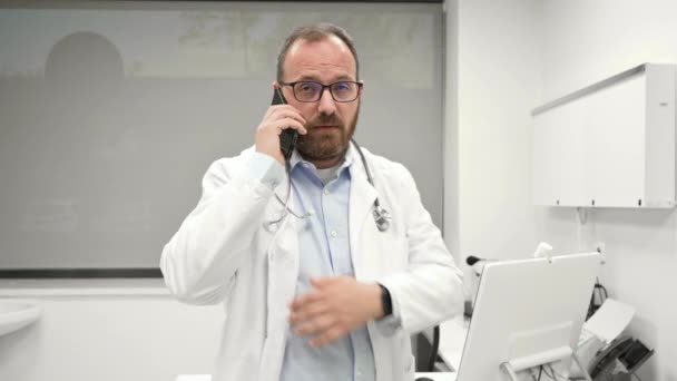 Doctor is talking on the cell phone by the window at a modern bright office background. — Vídeo de stock
