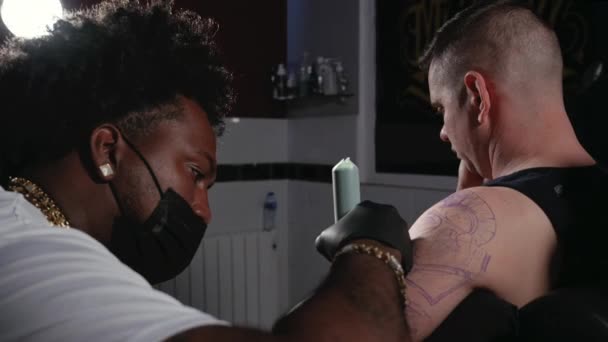 Tatuaje artista está realizando su trabajo en un joven en su estudio. Luz cinematográfica de un estudio de tatuajes. — Vídeos de Stock
