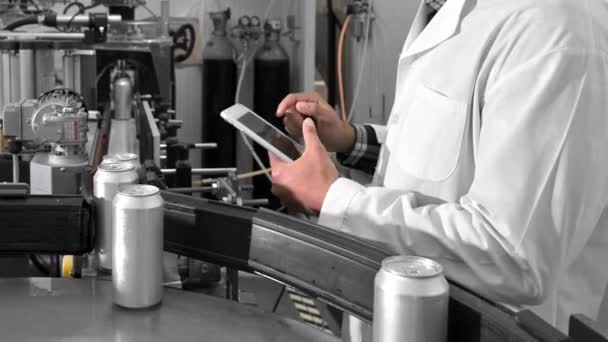 Type mannelijke werknemer op een tablet in de buurt van een transportband met blikken in de productielijn van de drankenfabriek, close-up. — Stockvideo
