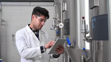 Industrial worker technologist in white coat and safety glasses looking at checklist and smiling at camera. Plant worker checking production in food industry.