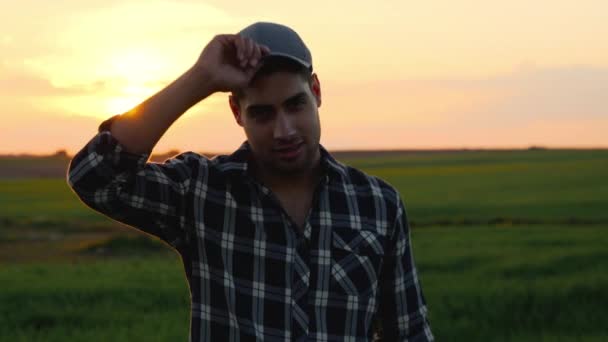 畑の中でカメラを見ている若い農民の肖像画と日没に笑みを浮かべて。農家は仕事の後に満足した彼の帽子を脱ぐ. — ストック動画