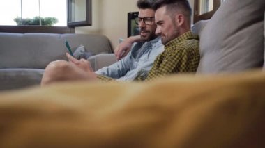 Happy young gay couple using mobile phone while sitting on a sofa in the living room. High quality 4k footage