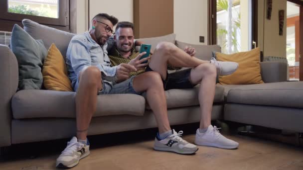 Happy Young Gay Couple Using Mobile Phone While Sitting Sofa — Stock Video