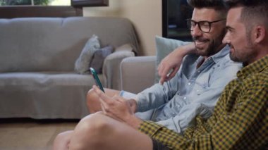 Happy young gay couple using mobile phone while sitting on a sofa in the living room. High quality 4k footage