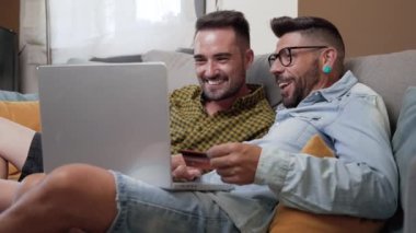 Happy young gay couple using laptop computer while sitting on a couch at home, shopping online with a credit card. High quality 4k footage