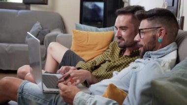 Happy young gay couple using laptop computer while sitting on a couch at home, shopping online with a credit card. High quality 4k footage