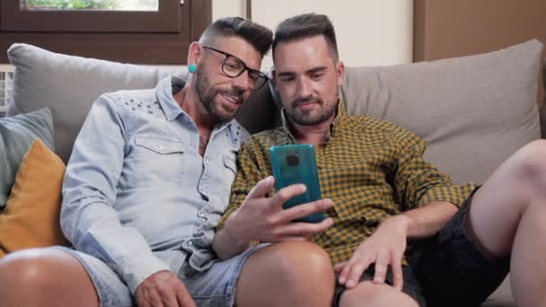 Весела Гомосексуальна Пара Сидить Дивані Обіймається Дивиться Мобільний Телефон Дивиться — стокове відео