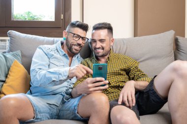 Oturma odasındaki koltukta otururken cep telefonu kullanan mutlu genç eşcinsel çift. Yüksek kaliteli fotoğrafçılık.