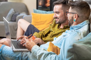 Mutlu genç eşcinsel çift evdeki bir kanepede oturup kredi kartıyla online alışveriş yaparken dizüstü bilgisayar kullanıyor. Yüksek kaliteli fotoğrafçılık