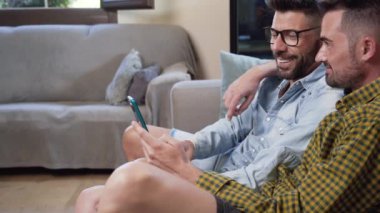 Happy young gay couple using mobile phone while sitting on a sofa in the living room. High quality 4k footage