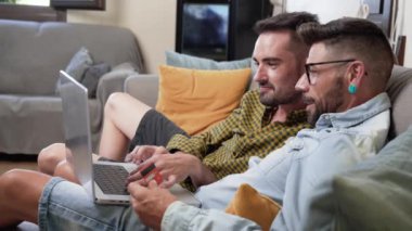 Happy young gay couple using laptop computer while sitting on a couch at home, shopping online with a credit card. High quality 4k footage