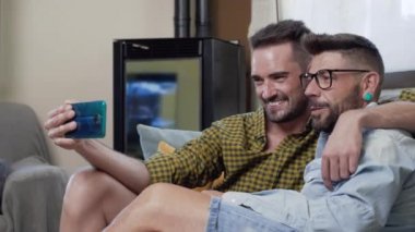 Gay Couple sitting in the sofa At Home Making Video Call On Mobile Phone. High quality 4k footage