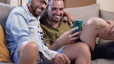 Cheerful gay couple sitting on the couch hugging and looking at the mobile phone, watching a movie, scrolling through the social network. High quality 4k footage