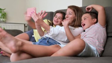 Happy mother with her kids are making a selfie or video call to father or relatives in a sofa. Concept of technology, new generation, family, connection, parenthood. High quality 4k footage